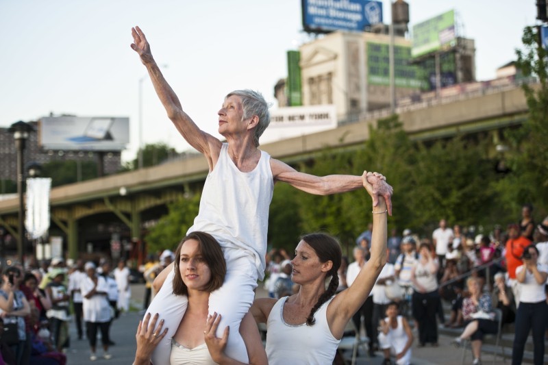 Two young women lift a more mature woman who wears a white tank top in a performance. The lifted dancer outstretches her right hand.