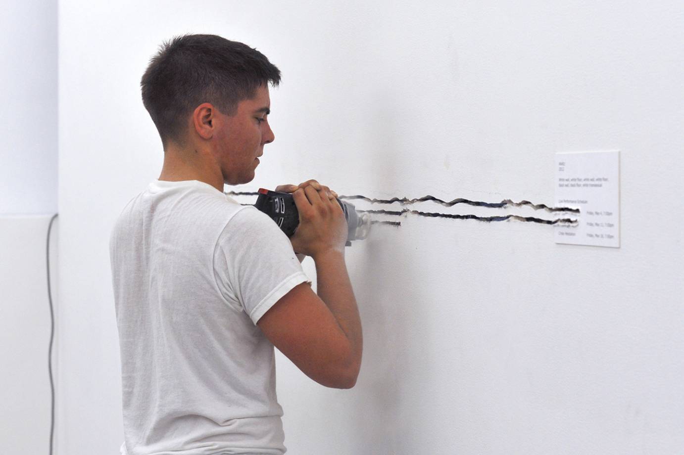 Yve Laris Cohen, dressed in a white t-shirt, creating deep lines in a white wall using a power tool.
