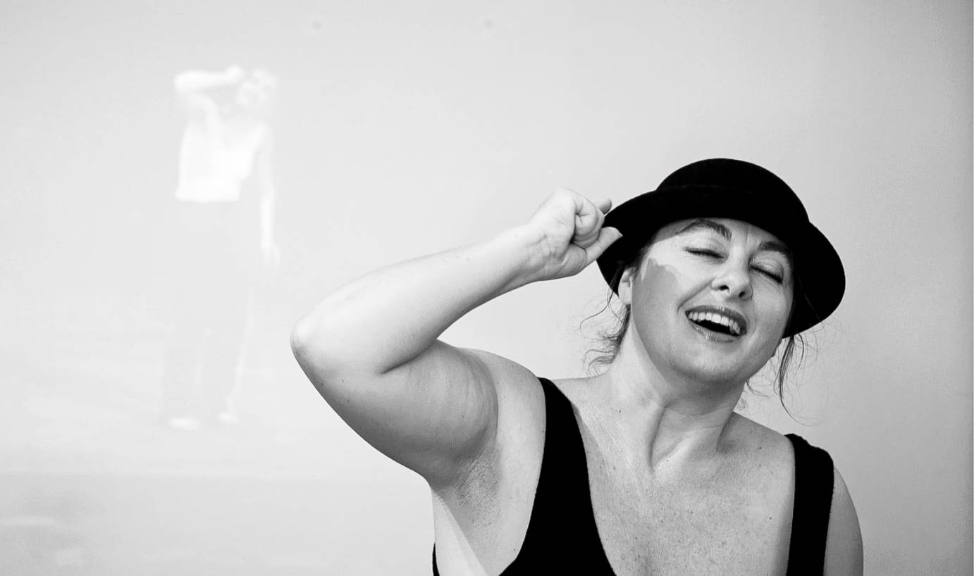 In a black fedora and tank top, Anabella Lenzu half closes her eyes while laughing