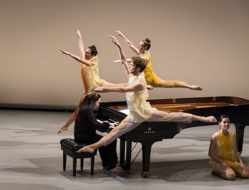 Three dancers in colorful gauzy tunics leap into the air around a piano. A pianist intently plays on. Another dancers kneels in front of the piano and looks out to the audience.