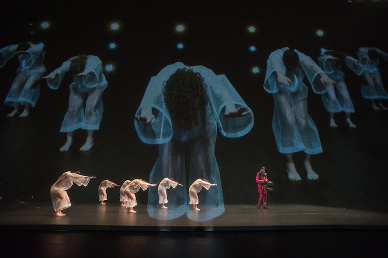 dancers in filmy white jumpsuits stand in side profile from the audience with their arms outstretched alongside their faces. A man in a red suit operates a steadicam to the right of them. They are projected larger-than-life on the scrim behind them.