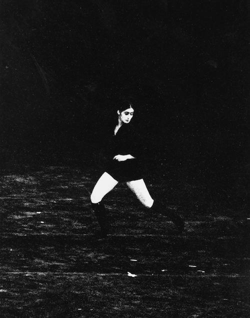 An archival photo of Twyla Tharp dancing in The Fugue in 1971. The photo is black and white and grainy. Tharp wears shorts and knee high boots