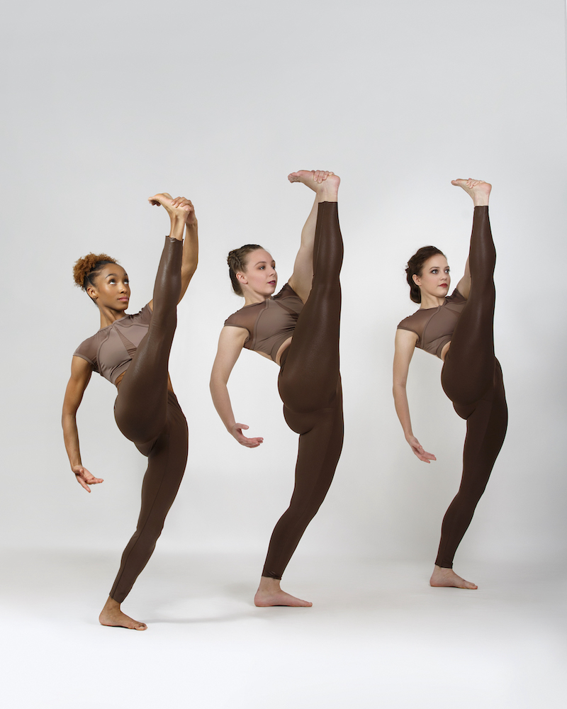 Three women  dancers of varieous complexions in brown unitard costumes. The top part is satiny brown with goeometric patterns and the bottom part are simple brown leggings. They are in unison holding their right leg high up in front of the with their left hand.