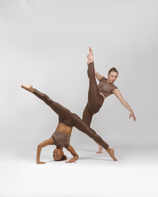 two female dancers one with brown skin and one with fair skin pose in highly technical balances. One balances upside down with weight distributed on her head, forearms and foot. the other balances in one leg on releve as she holds her other leg high in front of her. Both wear skin tight brown  brown costumes with bare midriffs 