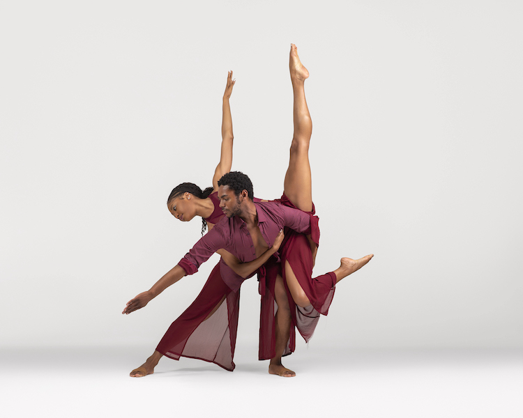 a male female couple. Both have light brown skin burgundy costumes with bottoms that are sheer and open to reveal leg. The woman balances on the man's back with her leg high in the air.