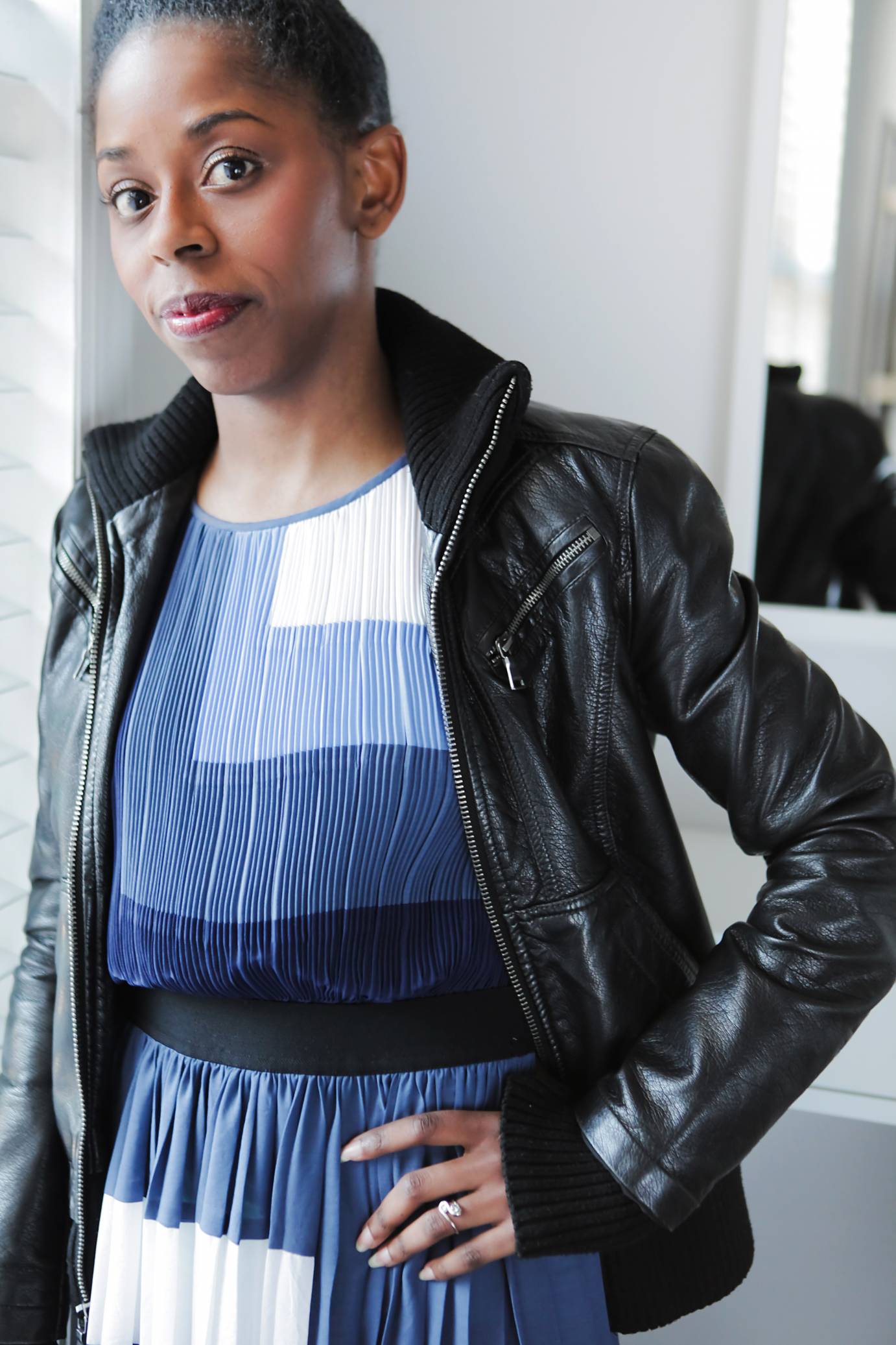 Portrait of Tiffany Rae Fisher a Black woman with big brown eyes and hair tightly pulled back leaning against a white wall as she stares out at us.She is wearing a dark blue, light blue and white pleated dress and a black leather bomber jacket.