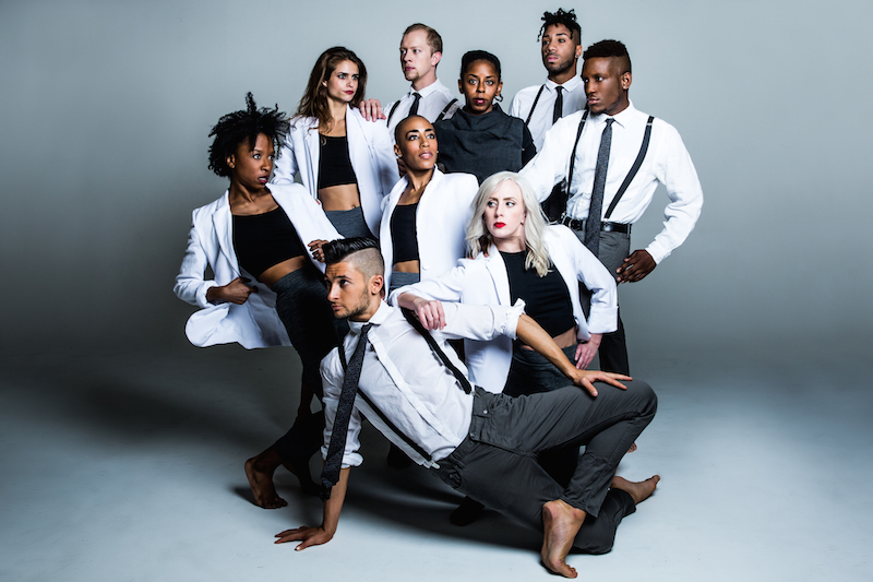 Dancers of Elisa Monte Dance wear black and white basics (blazers, jeans, button ups) and pose in a clump. Their gazes are in different directions. 