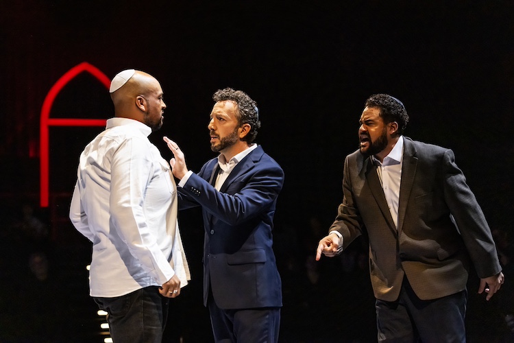 Three men wearing yarumukles that signify they are in a synagogue appear to be arguing. They are wearing everyday clothes ( not dance costumes). Two of them wear suits, not ties. The other is dressed more casually in a white top and dark jeans. To the left we see the red arch way symbolizing a house of worship.
