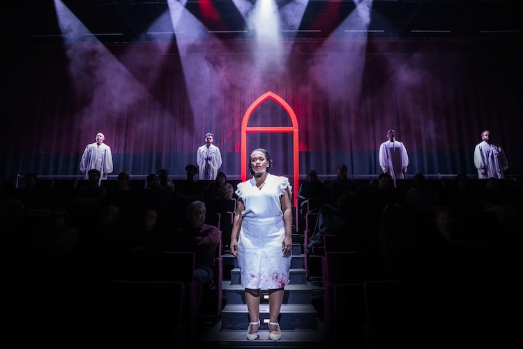 a Black woman in a white dress that is blood-stained on its bottom stands in front of a red lit archway that represents a house of worship. Four choir members in white robes stand behind her.
