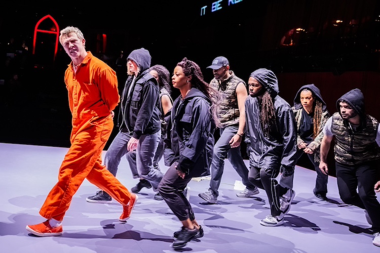 a white-colored man in an orange prison jump suit with matching shoes, leads a group of people in darker clothes, many in hoodies or caps, in a procession. He seems in the middle of saying something important as the others are quiet. 