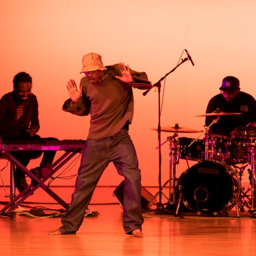 Man in cap with short brim, shoulders shrugged, arms bent with palms toward the audience, in loose dark clothing, dancing in front of keyboards and drums