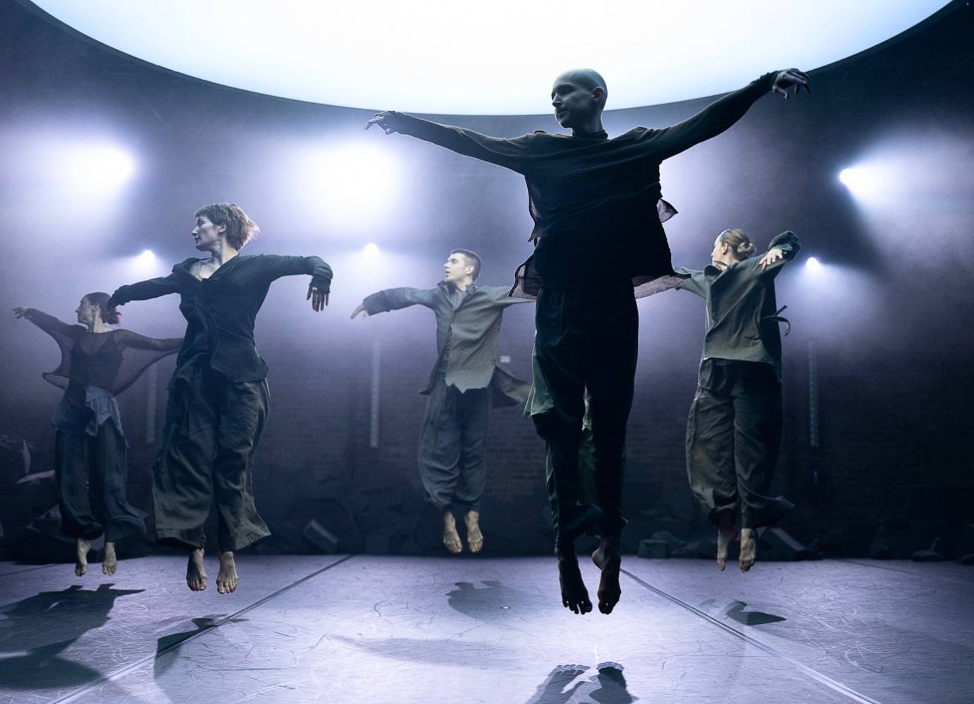 Fived dancers, three male and two female, face us and jump up.  Parallel legs point down and arms loosely fly sideways and upward, as the dancers look to their right. They all wear dark flowing shirts and wide-legged dark pants.