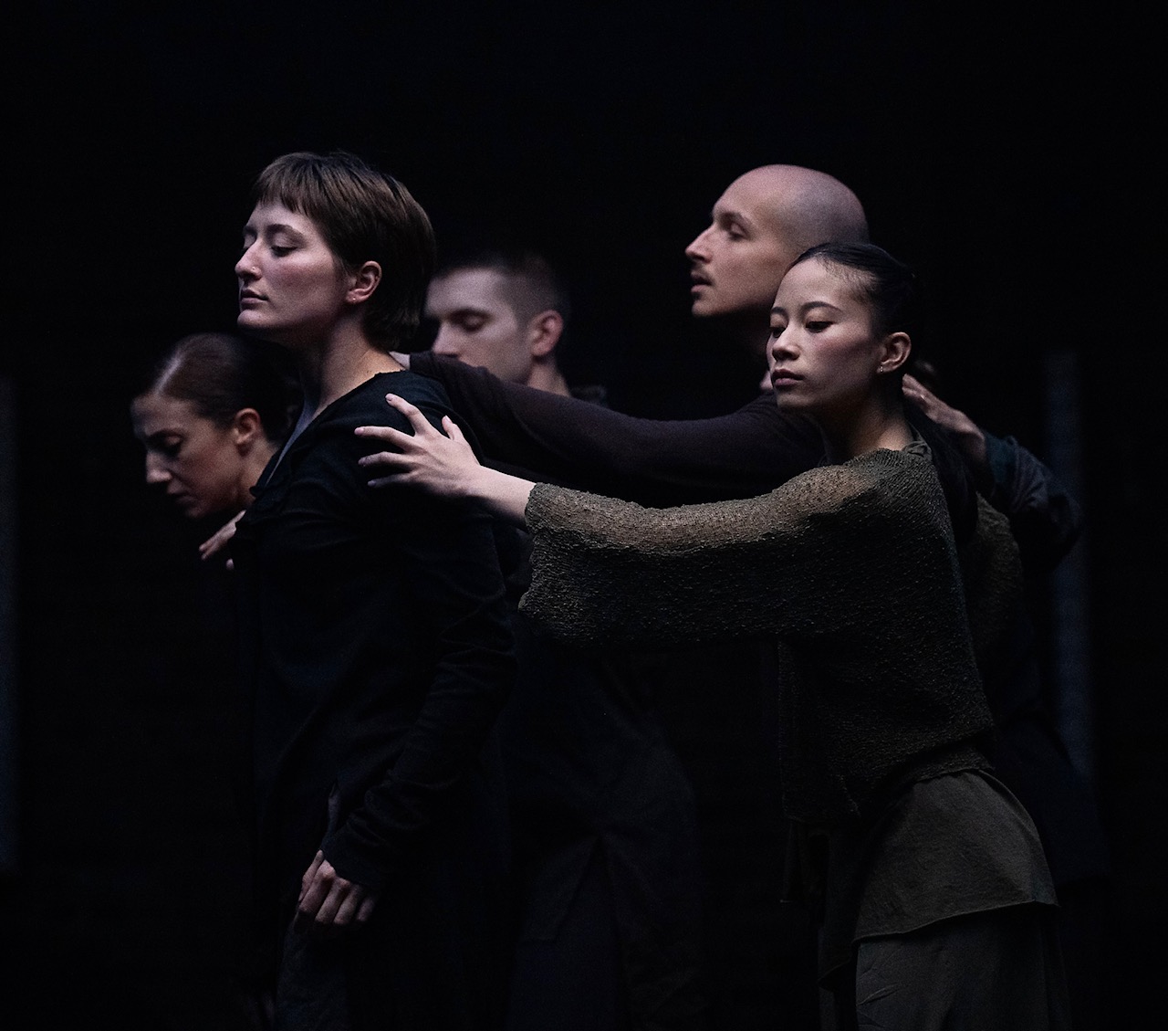 Five dancers' pictured in a head and torso shot tableau. Wearing dark colors so they melt into the black background we notice they face the right side, mostly with eyes closed, faces all poised in various states of reflection. 