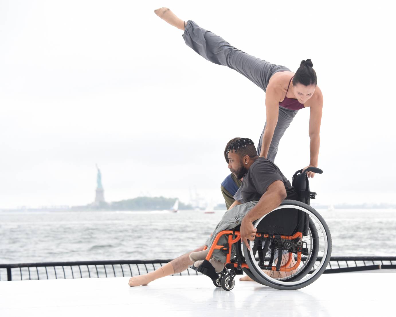A woman extends one leg sideways as she uses a wheelchair to elevate herself into the sky. The man in wheelchair leans slightly forward. The leg of another dancer peeks out by the wheel of  the wheelchair but the body remains hidden.