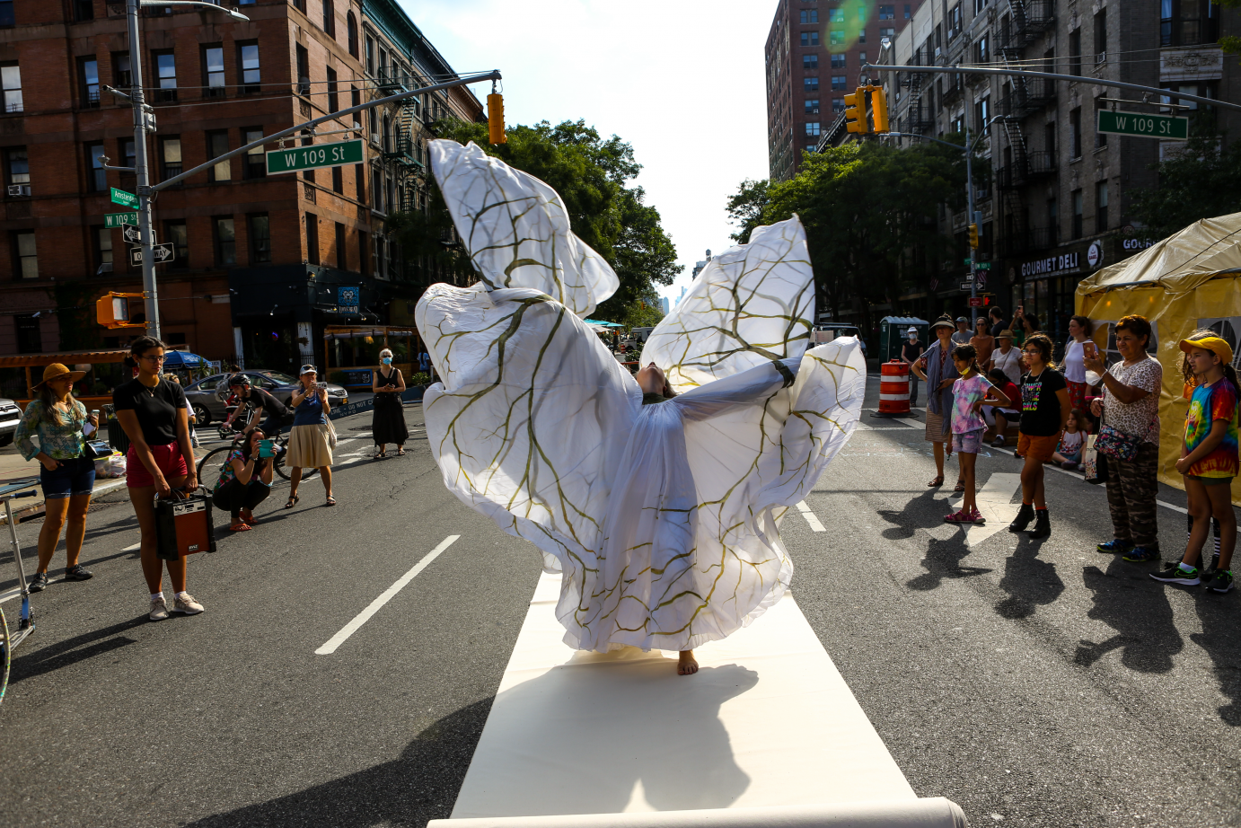 On a New York street, Jody Sperling wears a long white dress decorated with gold swirls. She lifts her arms in the air, head thrown back, as the fabric moves with her.