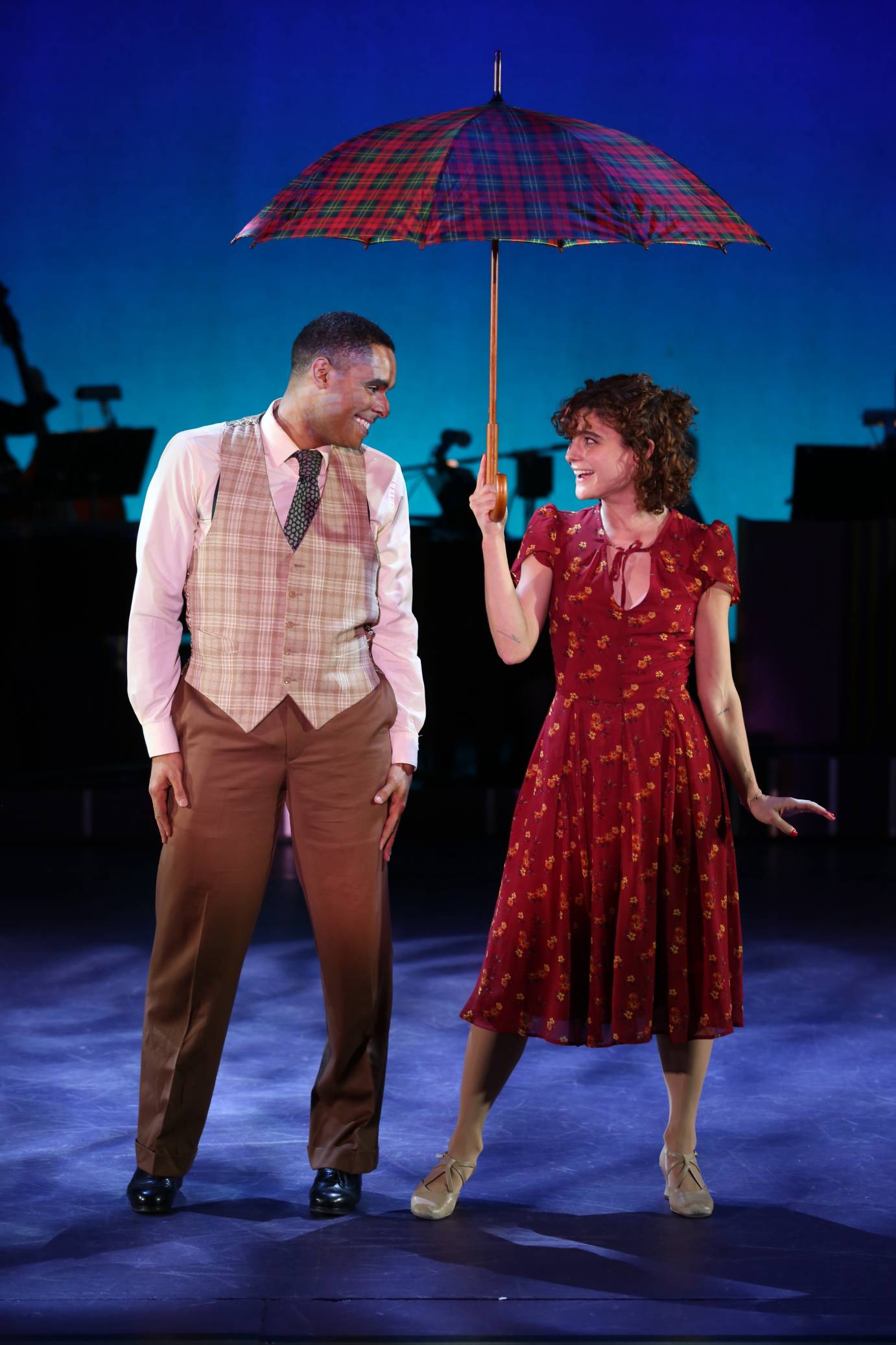 In a red printed dress, Melanie Moore holds an umbrella over a vest-clas man. Both are looking at each other, smiling.
