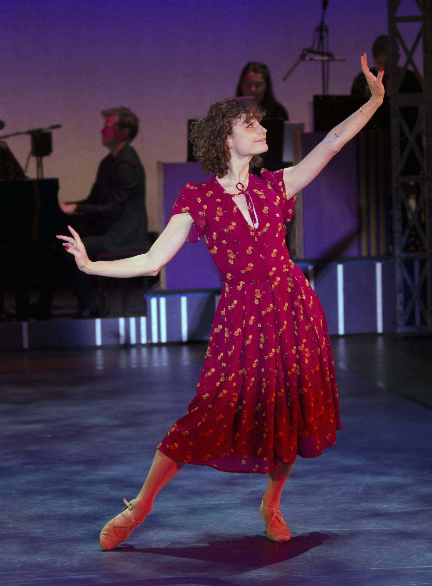 In a long maroon-printed dress, Melanie Moore holds her arms to the side, bent at the elbows and the wrists. She gazes upward to her extended arm, smiling slightly.