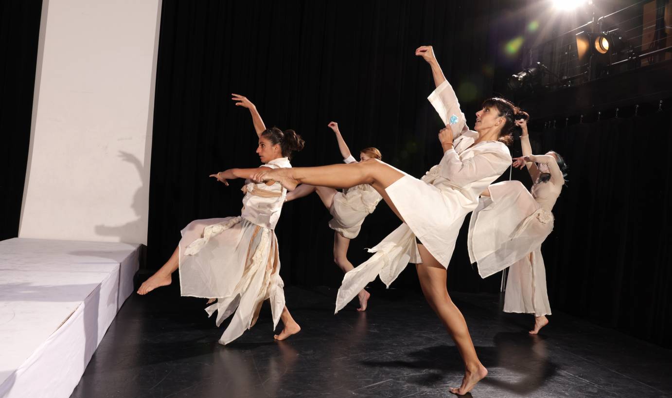A group of women in white lift a leg and arm while in profile. In the downstage dancer's free hand, she holds a blue lollipop.