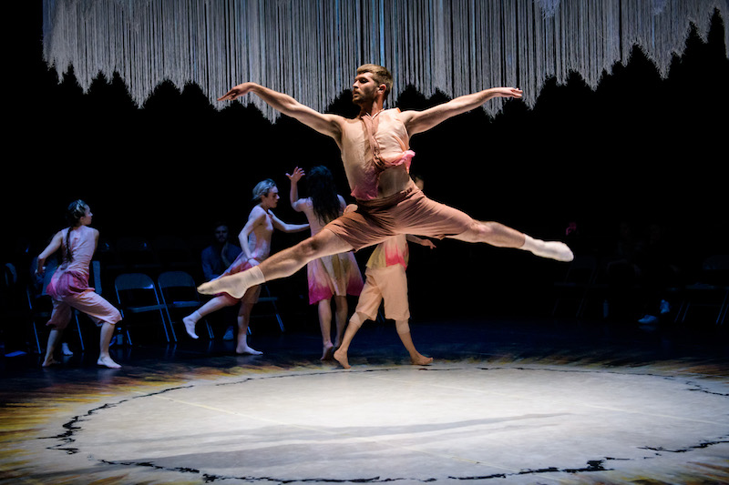 A male dancer in a pink hued gossamer costume leaps into the air. His legs extend perpendicular to the floor. Four other dancers move in the background. 