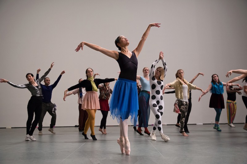 A woman in a blue skirt, black leotard and pointe shoes leads a group of MoMA employees in a dance