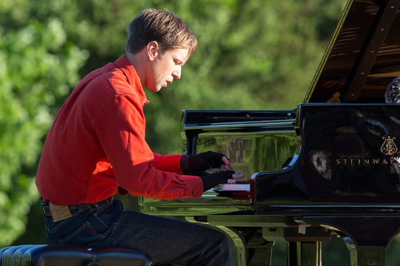 Hunter Noak wearing red shirt, blue denim pants, and black fingerless gloves, sits at a black Steinway grand piano playing it in Kaatsbaan park.