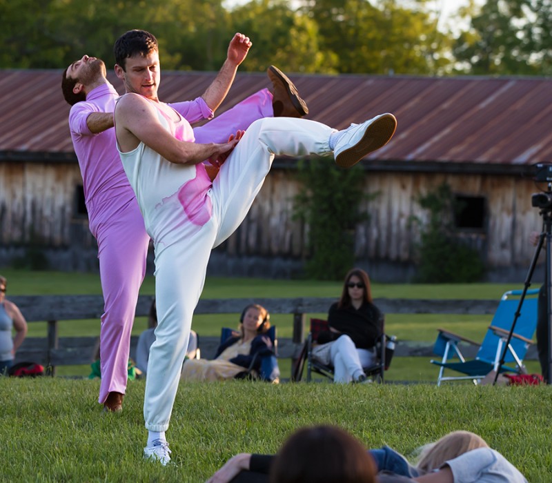 two male dancers perform in the grass. Each stands on one leg while the other leg is kicked high to their side. The man in the foreground is completely in white save for a pink stripe across his chest and his counterpart behind him is dressed completely in pink.