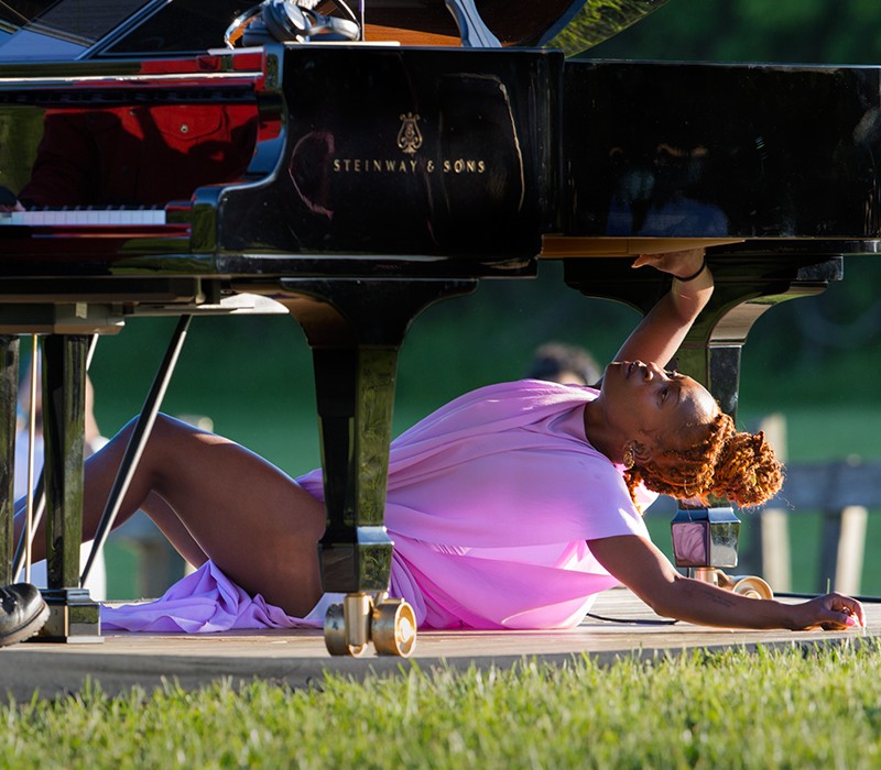 Coral Dolphin, the beautiful black woman dancer in the pink tunic, lays under the Steinway grand piano outdoors. 