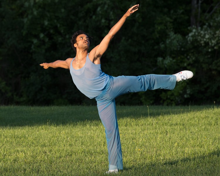 Taylor Stanley, dressed in sheer baby blue pants, and a sleeveless matching tank top holds a beautiful classic arabesque outside in the grass.