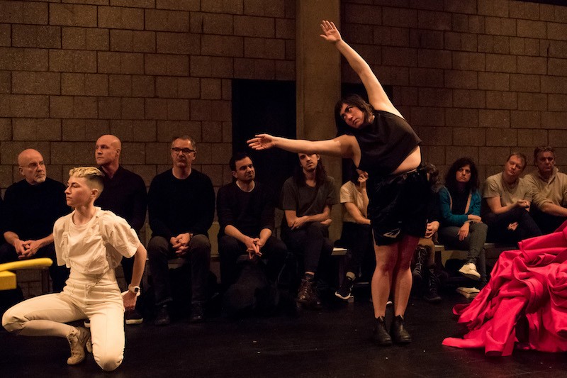 Jen Rosenblit in black boxing shorts and top reaches her arms over her head to the side. A performer in all white crouches to the right of her.