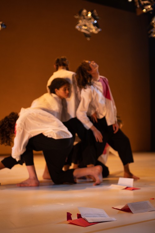 a quartet of teens huddle together facing different directions. they wear white button down shirts and black pants. Red journals surround them on the floor and silver mylar balloons float above.