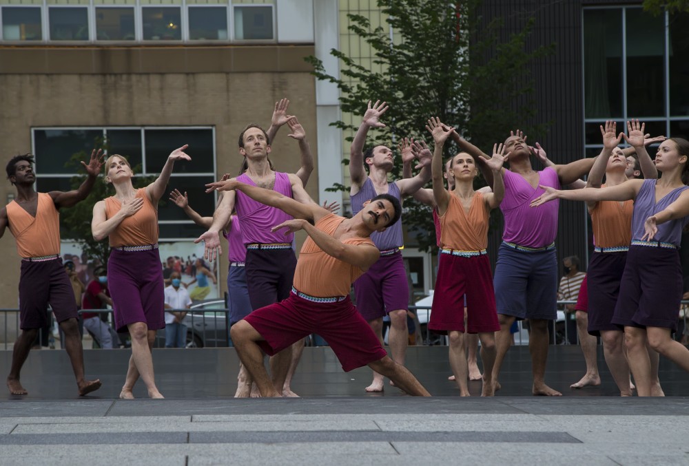Mark Morris group of dancers, approximately 12 people, women and men, of different skin colors, are outdoors in a cement plaza, dancing with arms outstretched. They are wearing similar costumes dark shorts and light v-necked sleevless tops in shades of purple and orange