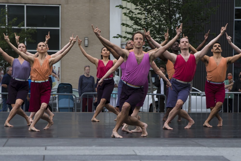 the 12 or so dancers all wearing shorts of various shades of purple and magenta , with sleevless tops of purple and magenta, facing us with their arms above them in a u-shape.