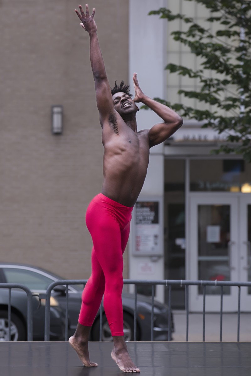 a black man in red tights with a pained expression on his face extends one are up into the sky and the other is bent, palm facing the side of his face and outstretched. Extreme emotion is displayed.