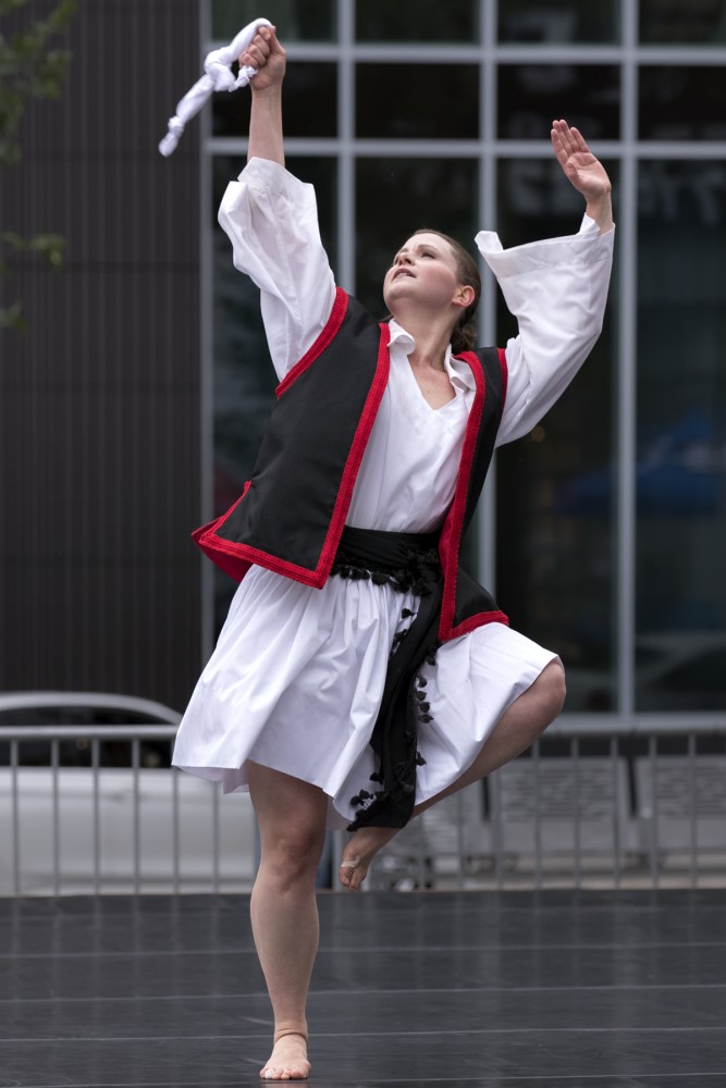 a woman in a Greek folk cosutme of mainly white with a black vest and red trim, looks to her upward right corner her arms extended above her, one of her legs bent in the air as she balances on the other