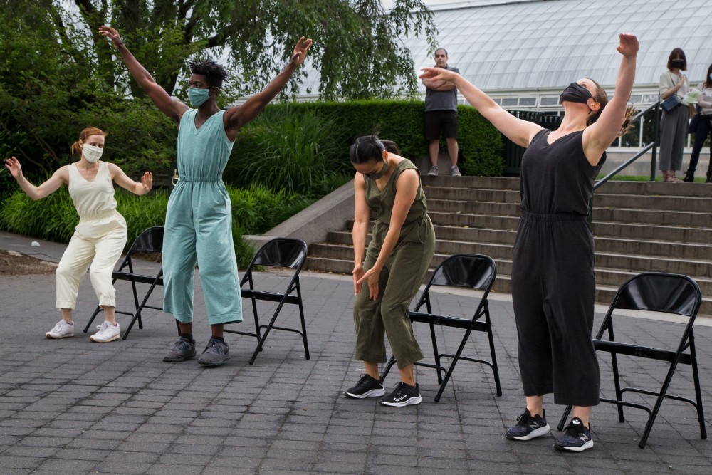 Four dancers of various skin tones are in various stages of rising from fold out chairs.They are performing outside in the garden wearing pantsuits of yellow, aqua, olive, and black with masks to match. They wear sneakers. 