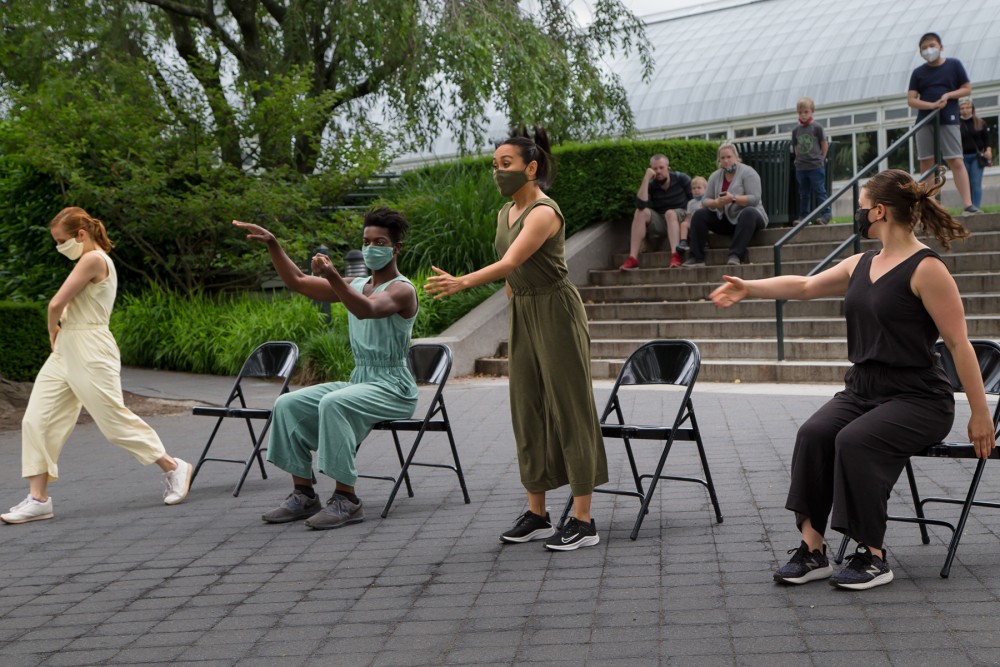 four dancers in pastel and dark jumpsuits, and sneakers, wearing masks that match their clothes...in different stages of sitting and rising from fold out chairs