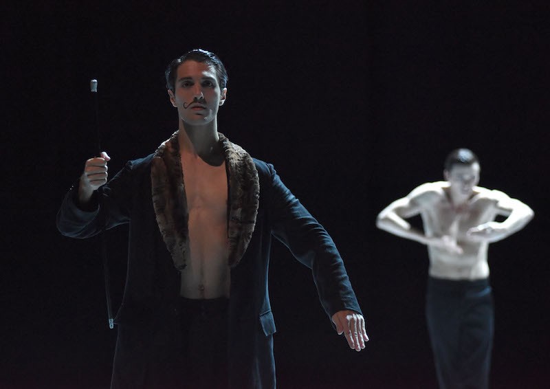 A man with a mustache stand with a cane and another man, who is bare chested - dances in the background