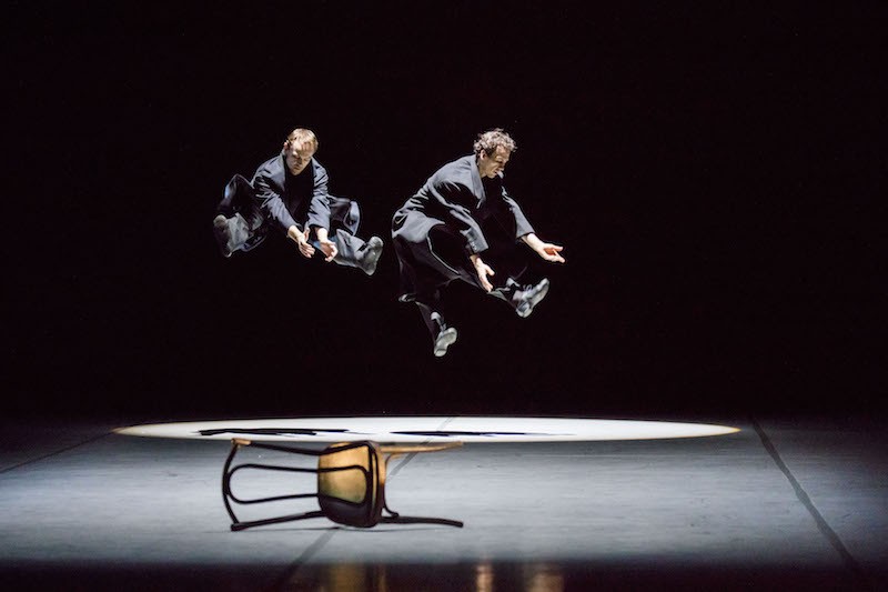 Two men jump with their legs outstretched into the air. Chairs lay on the ground.