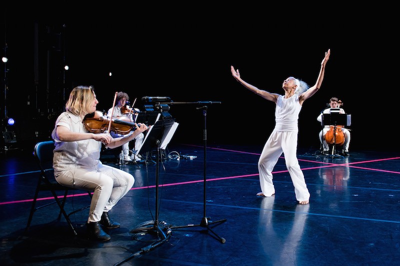 McIntyre dressed in white reaching her arms overhead while the string trio sits in chairs around her