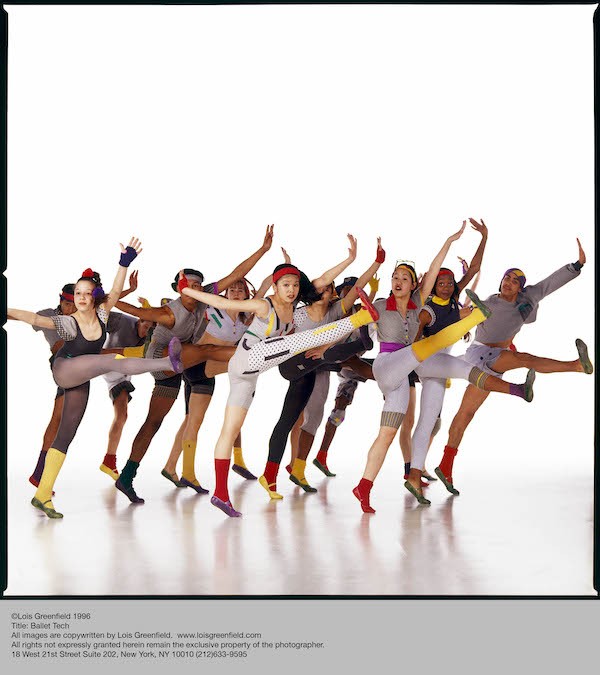 A group of Ballet Tech students in colorful leotards, tights, shoes and leggings, lift their right leg and flex their foot. They look out at the camera