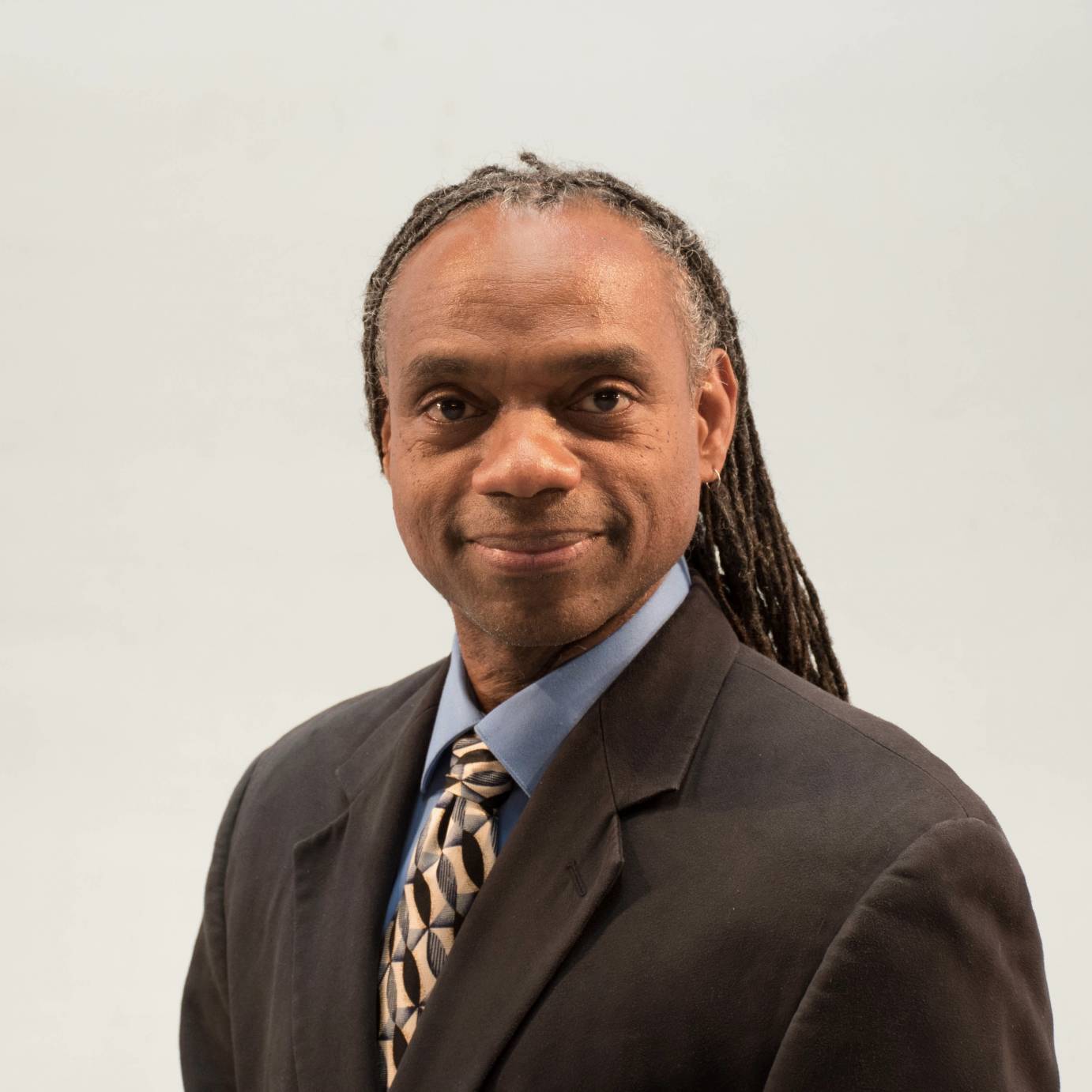 Professor Paul Anthony Dennis, an African American male with greying dreadlocks, is wearing a smart suit with a blue collared shirt and a patterned neck tie. 