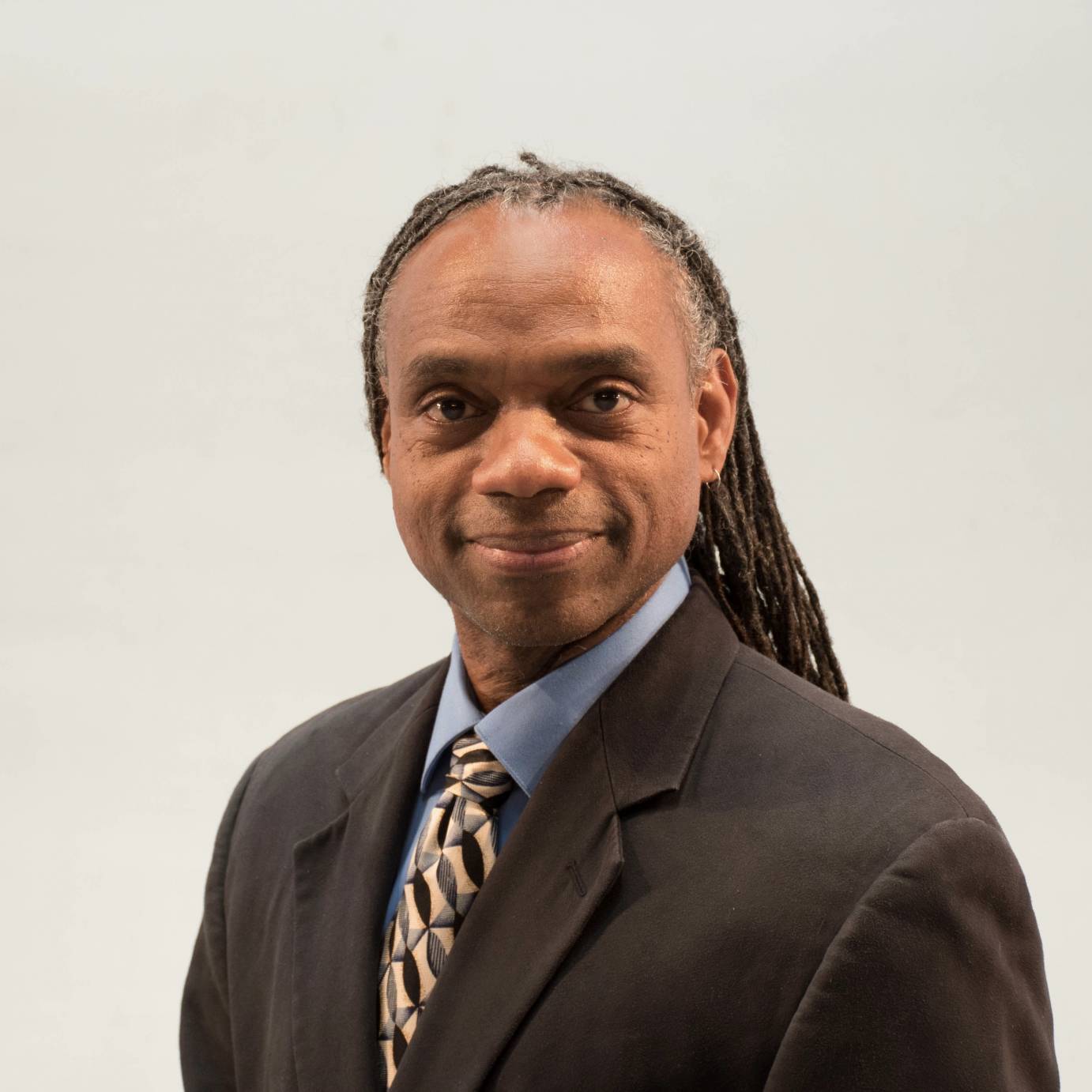 Professor Paul Anthony Dennis, an African American male with greying dreadlocks, is wearing a smart suit with a blue collared shirt and a patterned neck tie.