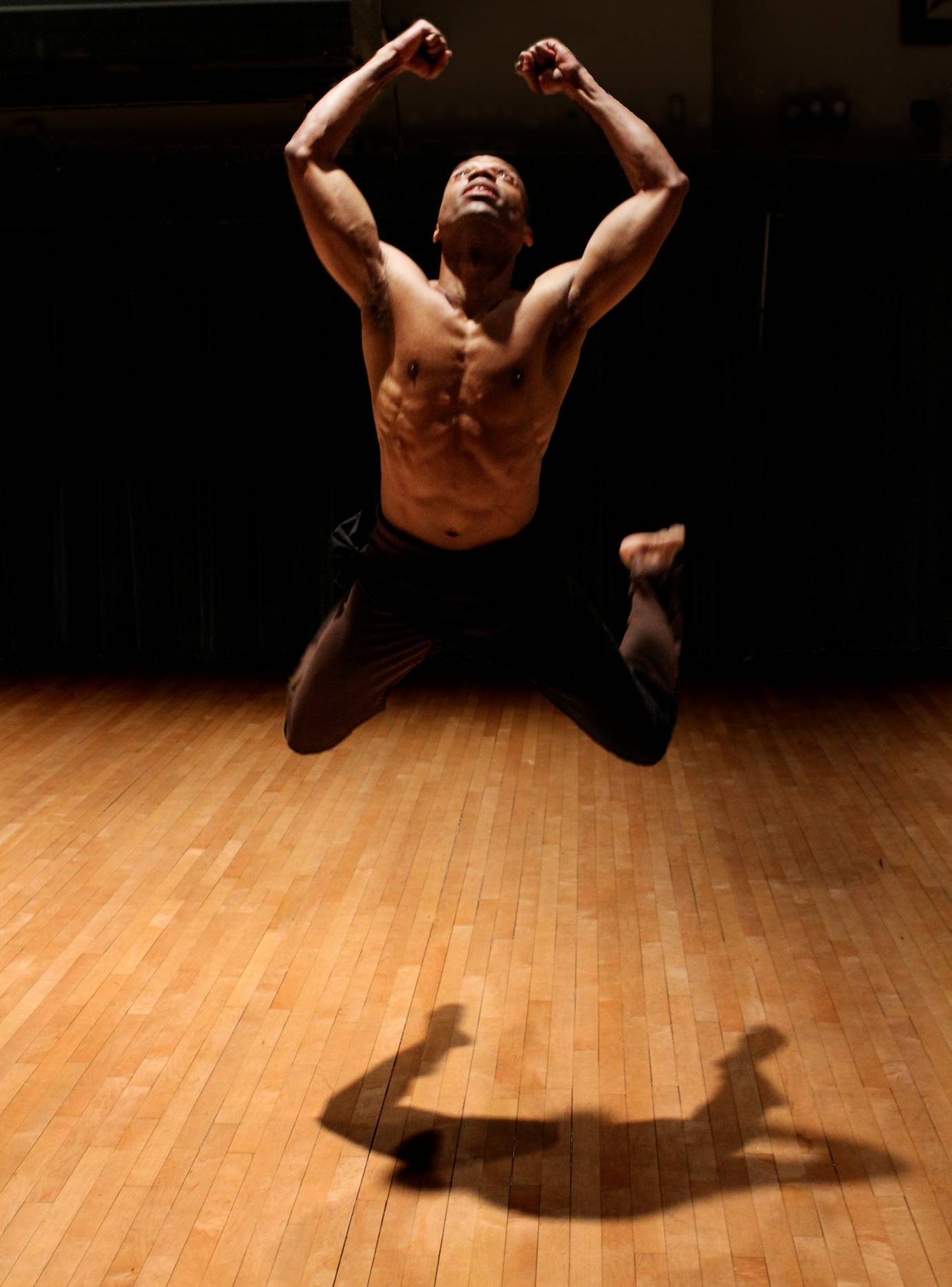 Paul Anthony Dennis, who is topless with black pants, leaps and is completely suspended in the air, his knees bent at an angle.