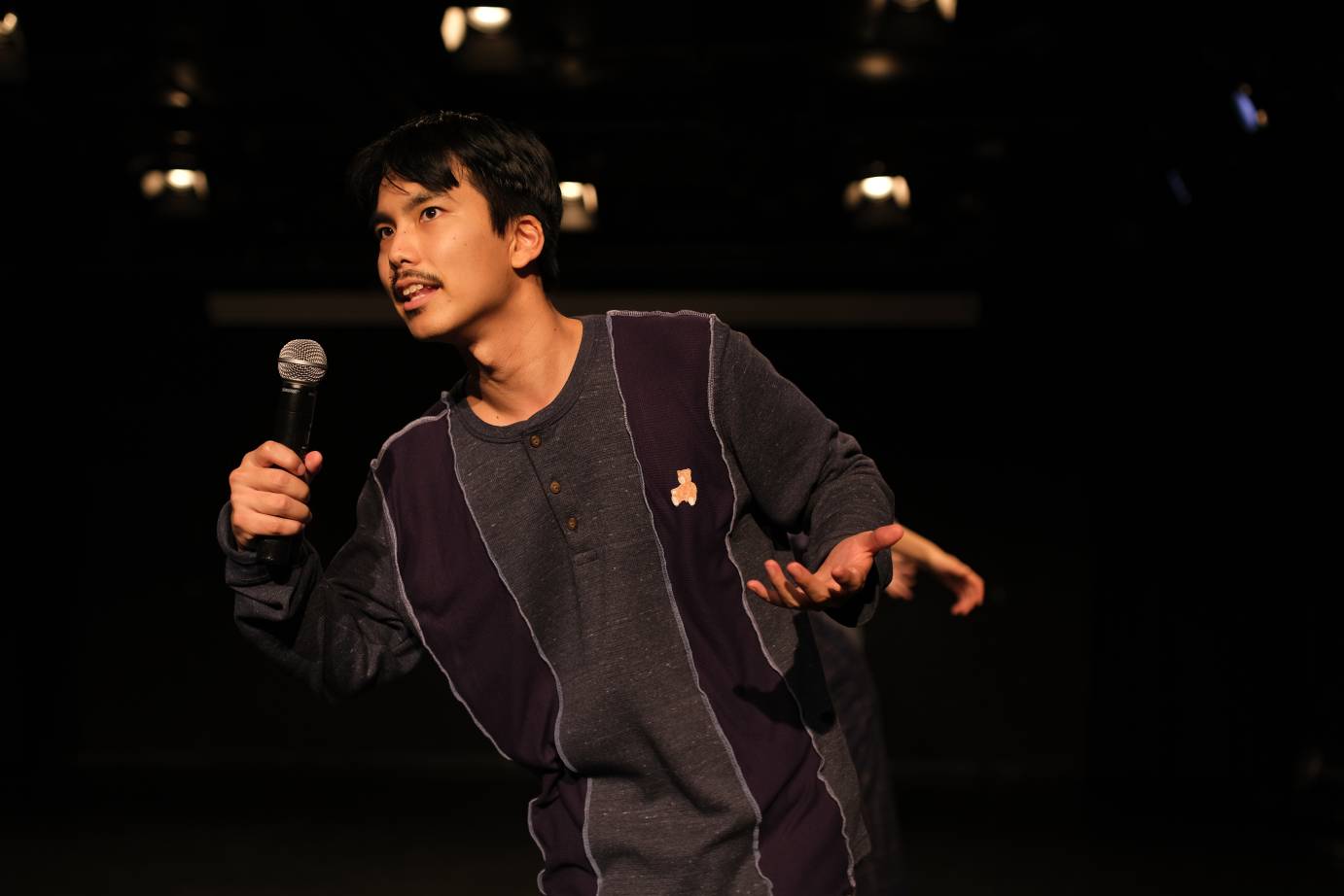 A mustachioed man speaks into a microphone wearing a dark costume against a black background.