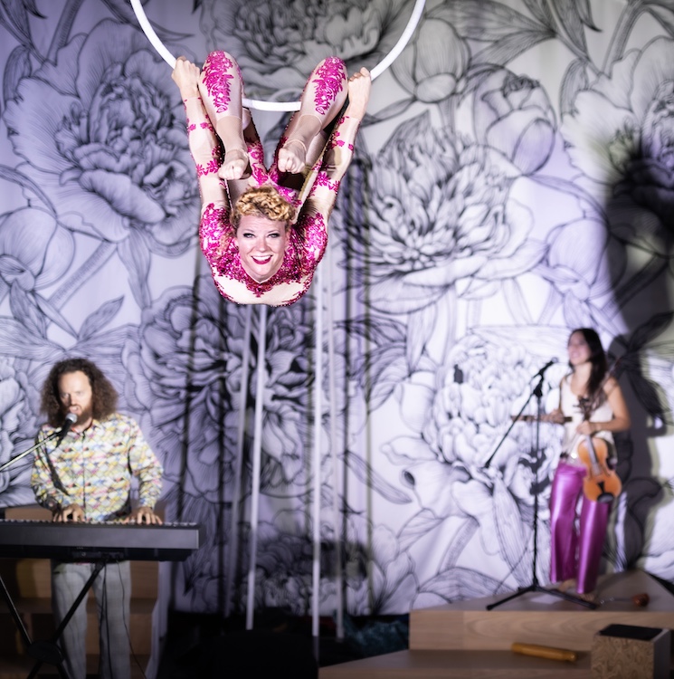 against the floral blk and white backdrop, Ellie Steingraeber, hangs in the air, her legs curling around a large suspended white hula hoop, the musicians play in the background