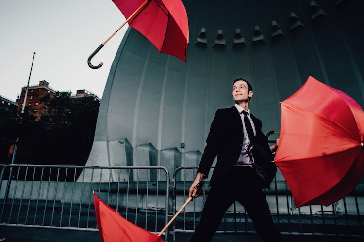 a press photo of Kyle Driggs wearing a black suite , looking dapper, as he juggles three large red umbrellas
