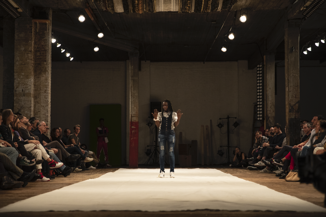 A girl in overalls stands with her arms bent and up at the elbows