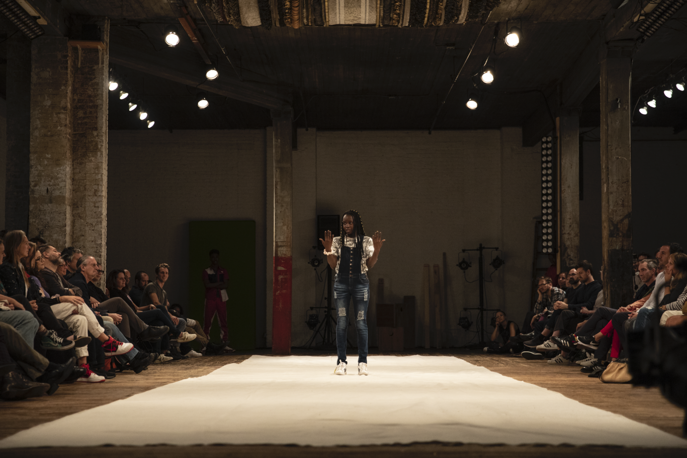 A girl in overalls stands with her arms bent and up at the elbows 