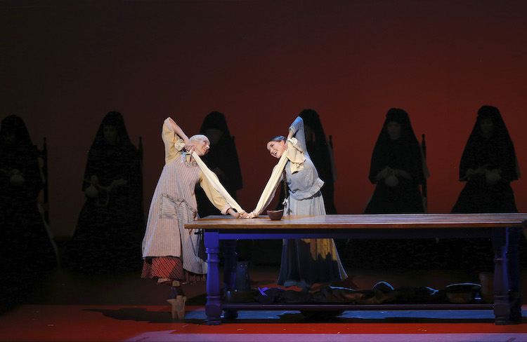 Luciana Parri and Cassandra Trenary, garbed in kitchen clothing, lean towards each other smiling as the strech their dough on the kitchen table... Behind them are a chorus of  silouhetted black figures in rocking chairs.