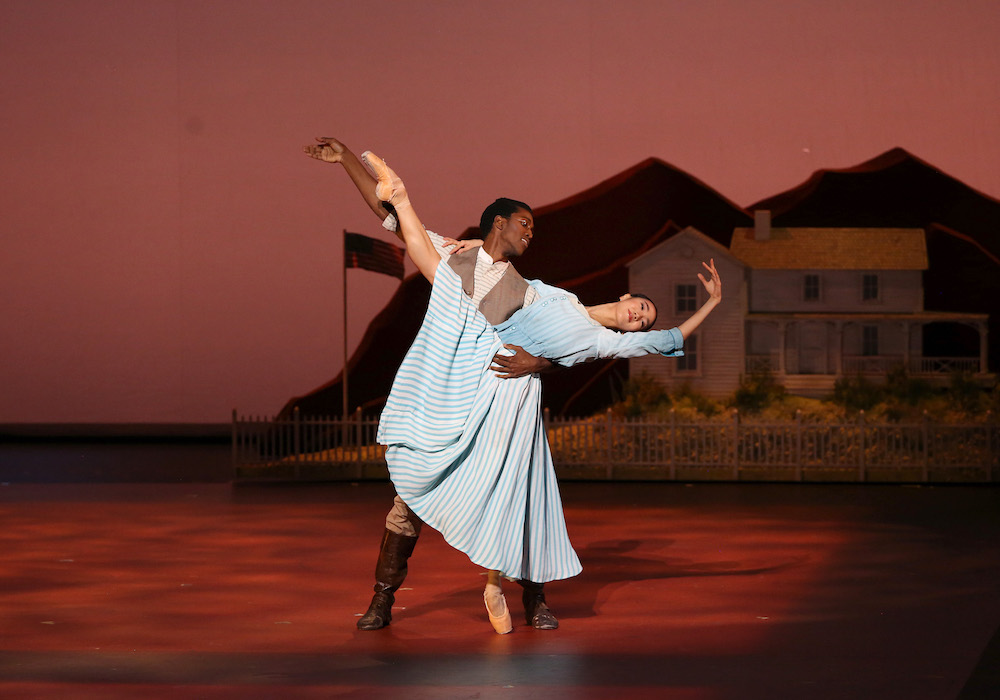 Calvin Royal grabs SunMi Park by her waist as she leans swooning in a back bend with one leg and pointed foot gesturing toward the sky. She is dressed in a long full dress of blue he wears brown with a white shirt and behind then is a set of houses. 